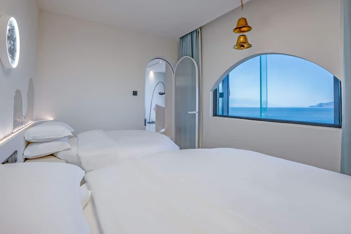 Una habitación de hotel moderna con una cama grande, un ventanal con vista al mar y una lámpara de pared.