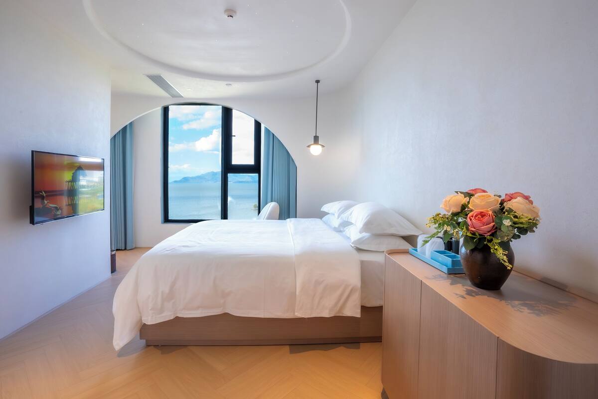 Un dormitorio moderno con un gran ventanal, una cama, un televisor y un jarrón con flores.