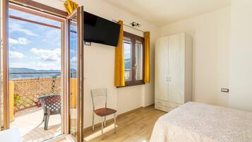 A bedroom with a bed, a chair, a wardrobe, a TV, and a sliding glass door leading to a balcony.