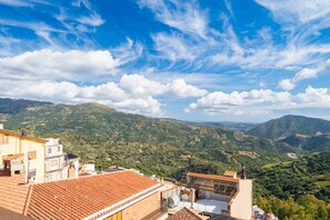 A view of a mountainous landscape with terraced vineyards, traditional buildings with red-tiled roofs, and a clear blue sky with scattered clouds.