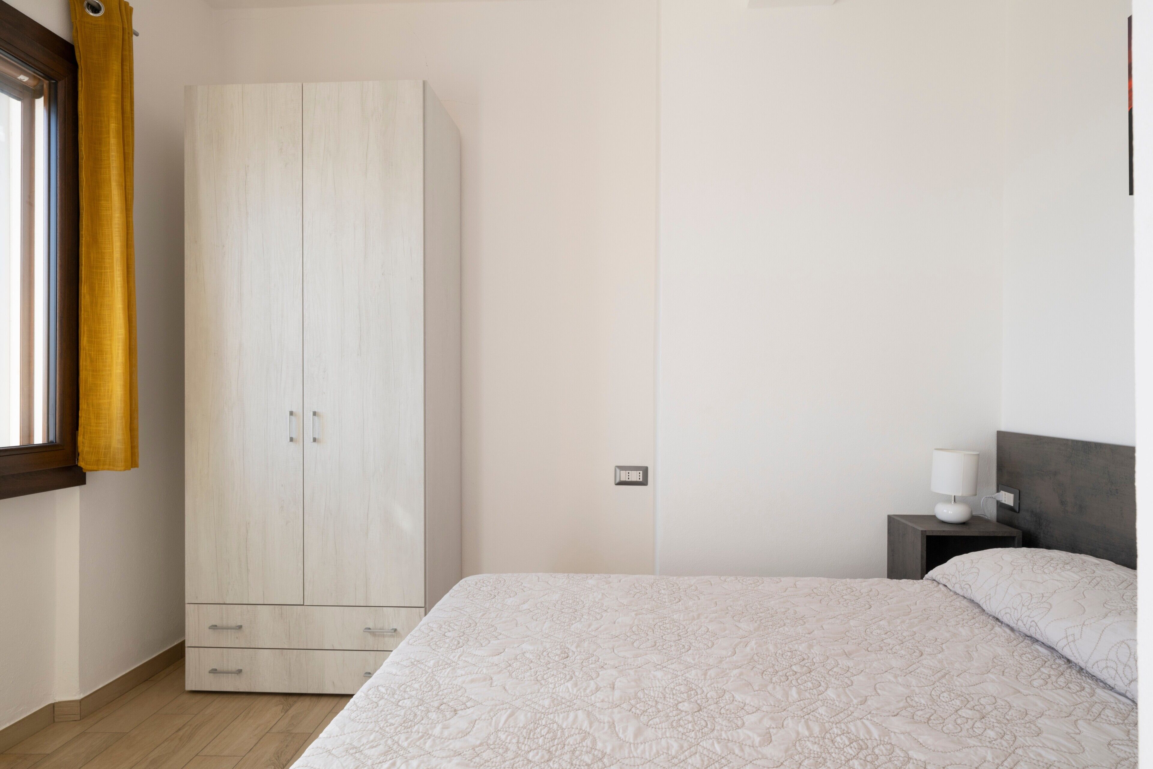 A bedroom with a bed, a wardrobe, a nightstand with a lamp, and a window with curtains.