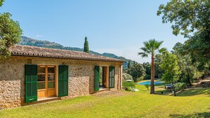 A stone building with green shutters, a grassy yard, and a pool in the background. - Villa 'Can Perot' with Mountain View, Private Pool and Wi-Fi (Pollença)