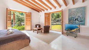 A spacious room with a bed, a TV, a chair, and a table. There are large windows with a view of greenery. - Villa 'Can Perot' with Mountain View, Private Pool and Wi-Fi (Pollença)