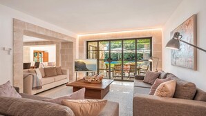 A modern living room with a large window, a flat-screen TV, and a dining area with outdoor seating. - Villa 'Can Perot' with Mountain View, Private Pool and Wi-Fi (Pollença)