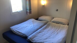 Une petite chambre d’hôtel, composée d’une seule pièce, avec deux lits, un banc, une fenêtre avec des rideaux et un luminaire fixé au mur.