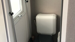 Une petite salle de bain étroite, avec des toilettes, une fenêtre et un balai appuyé contre le mur.