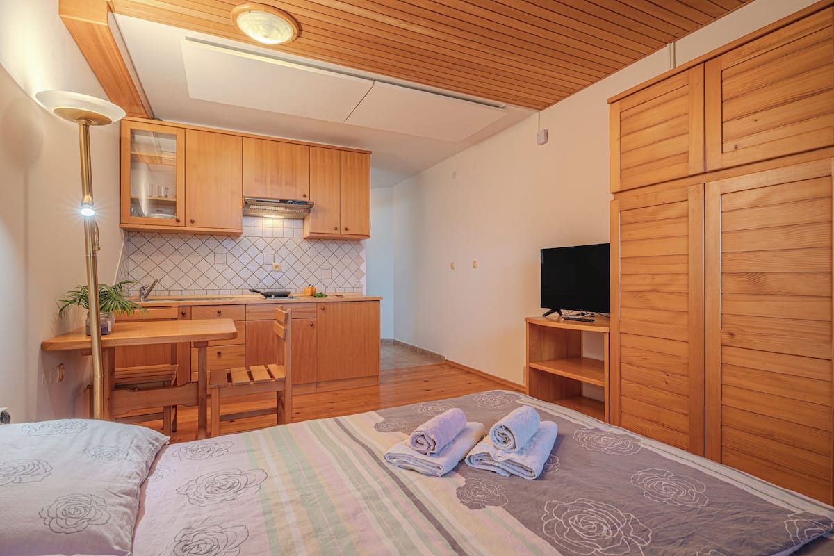 A compact room with a bed, a desk, a kitchenette, and wooden cabinets.