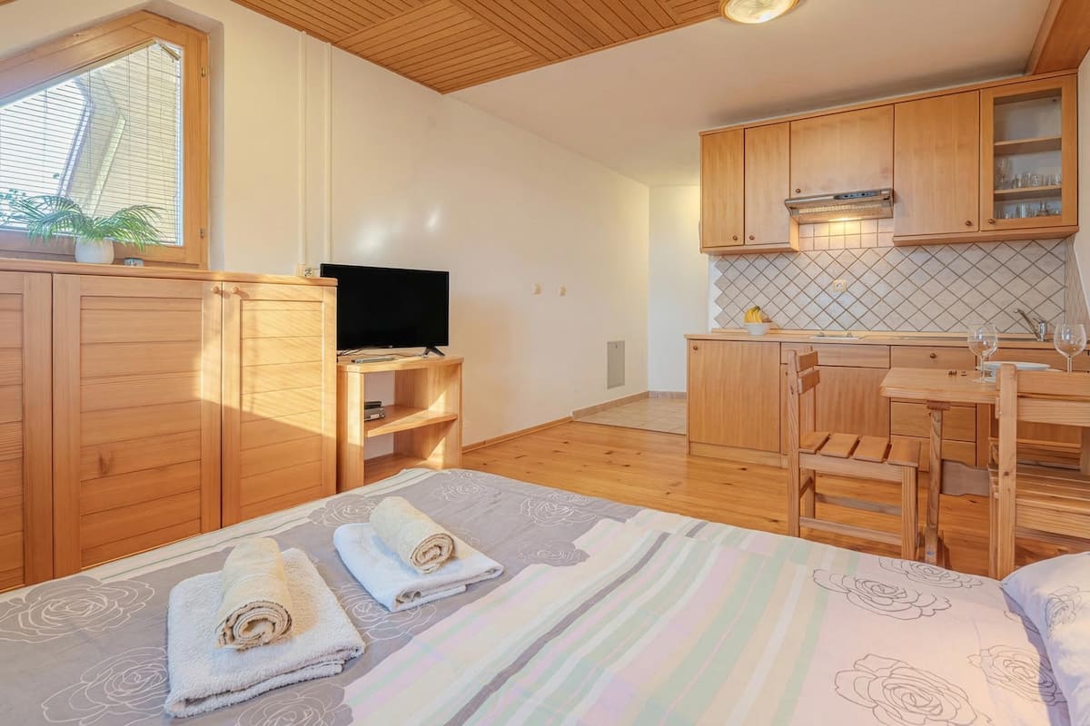 A compact room with a bed, a TV, a small kitchen area, and wooden cabinets.