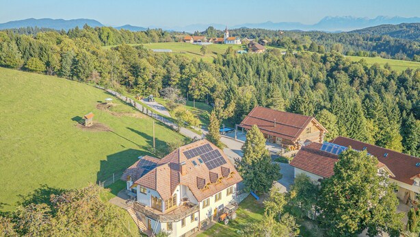 A residential area with houses featuring solar panels, surrounded by lush greenery and mountains in the background.