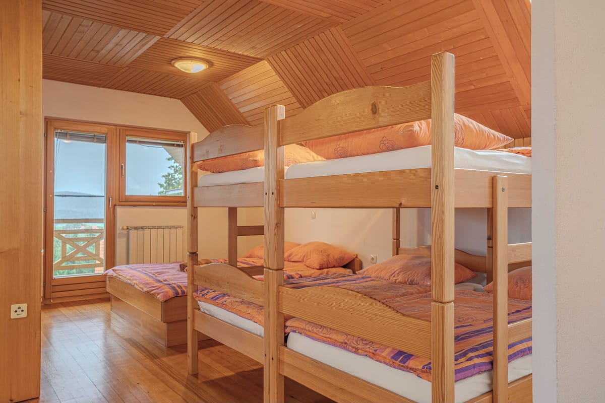 A room with three bunk beds, wooden flooring, a sloped ceiling with wooden beams, and a window with a view of the outdoors.