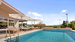 A swimming pool with lounge chairs and umbrellas, surrounded by a stone wall and vegetation.