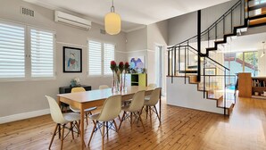 Un vaste espace dédié à la salle à manger, avec une table et des chaises en bois, un escalier moderne et de grandes fenêtres équipées de stores.
