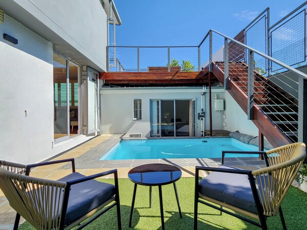 A modern outdoor pool area with a wooden deck, two lounge chairs, a small table, and a staircase leading to a second floor.