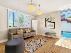 A living room with a sofa, ottoman, and rug, large windows with blinds, a ceiling light, and artwork on the wall.