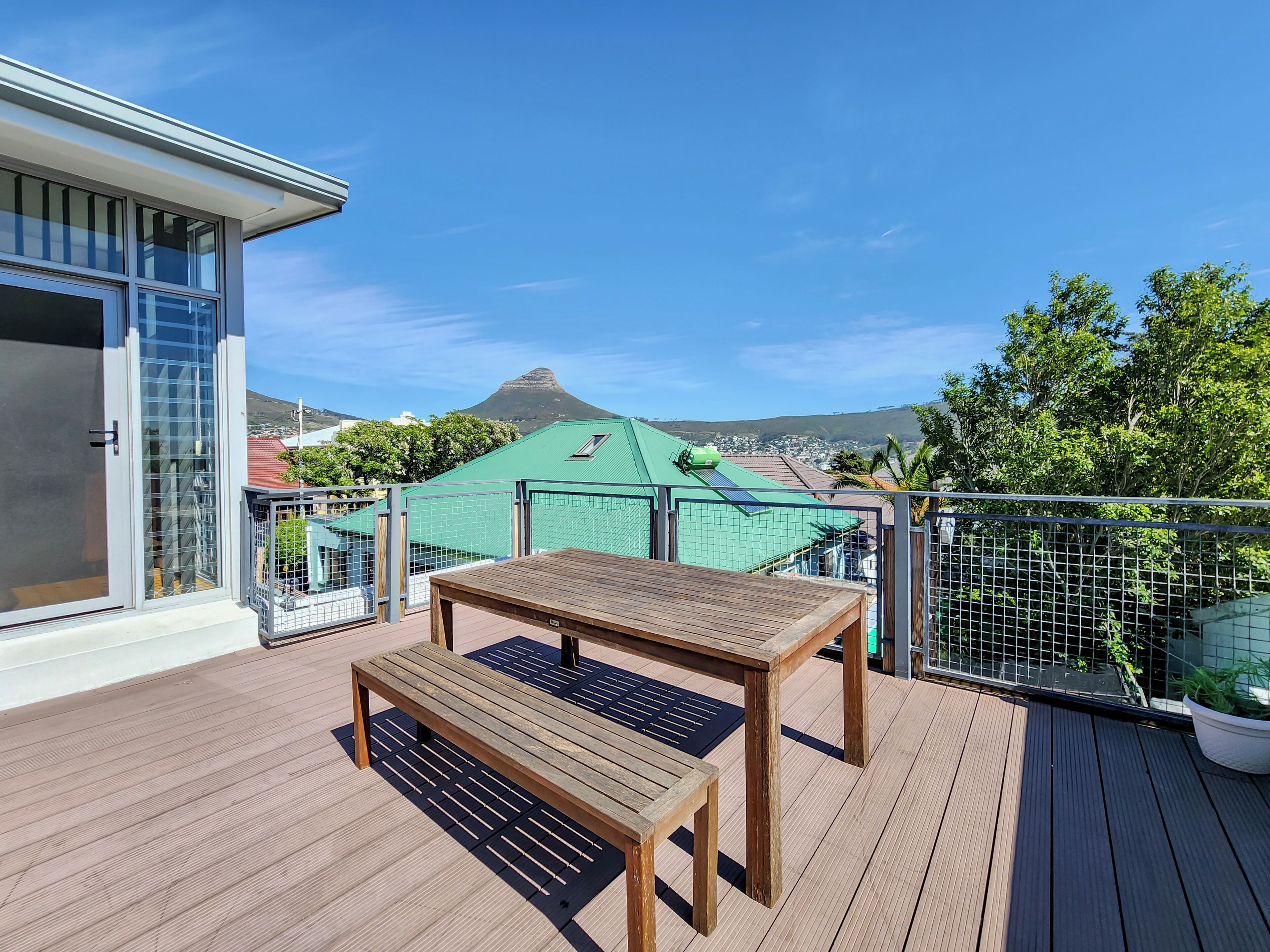 Una terraza de madera con una mesa, bancos, una puerta de cristal y vistas a un edificio con techo verde y una montaña a lo lejos.