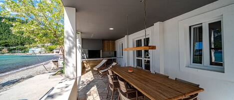 Un área de comedor al aire libre con mesa y sillas de madera, un lámpara colgante y vistas a una piscina y árboles.