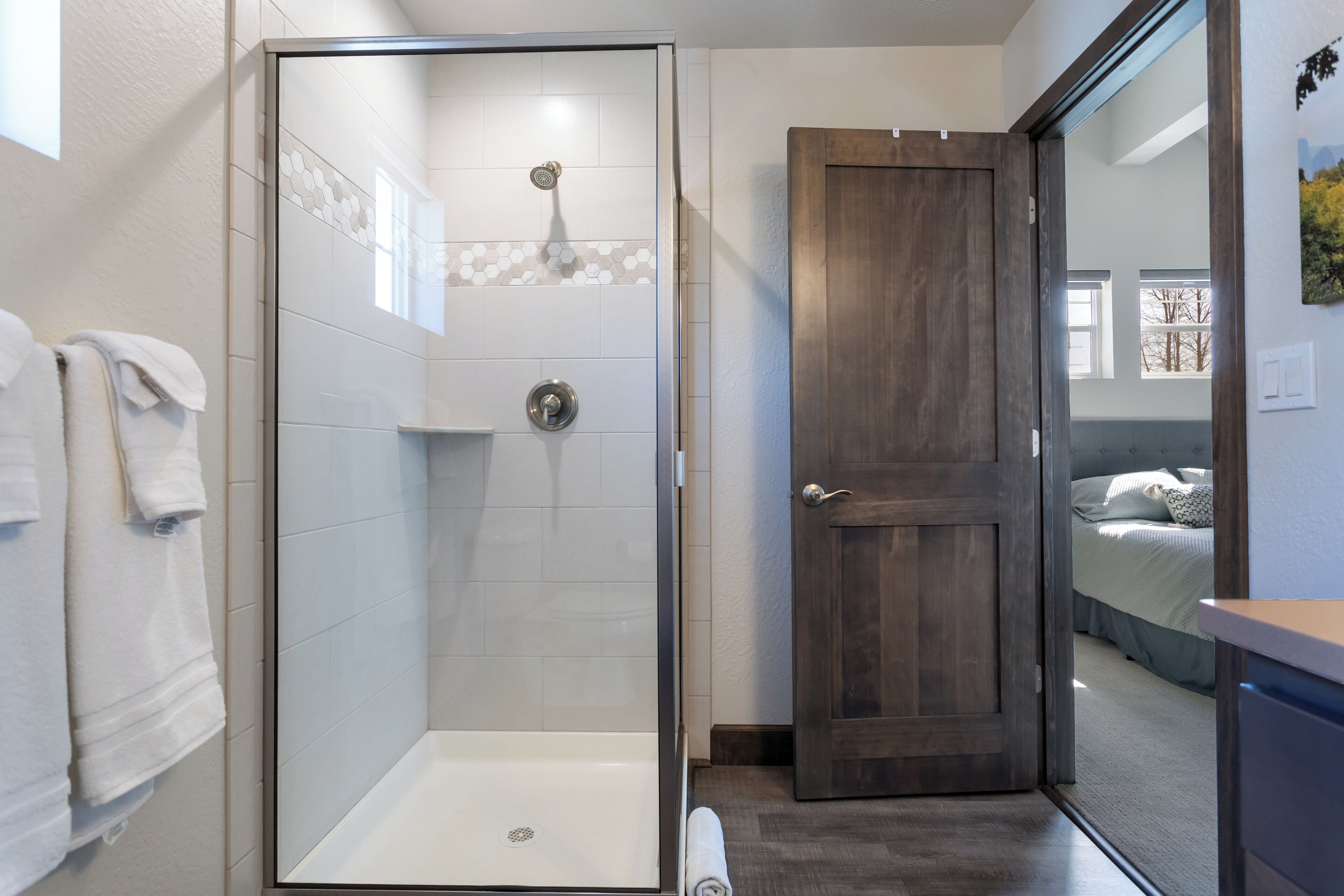 A modern bathroom with a glass-enclosed shower, a wooden door, and a visible bedroom with a bed and window.
