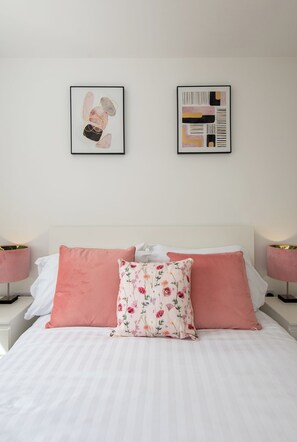 Ein ordentlich bezogenes Bett mit rosa-weißer Bettwäsche, zwei gerahmte Kunstwerke darüber und Nachttischlampen auf beiden Seiten.