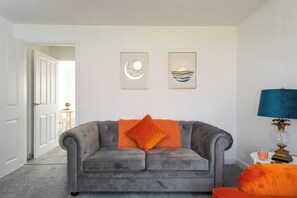 Um sofá cinzento com uma almofada laranja, uma luminária azul e duas obras de arte emolduradas na parede.