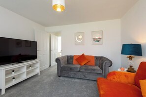 Ein Wohnzimmer mit Flachbildfernseher, grauem Sofa, orangenen Kissen, einem weißen Unterhaltungsschrank und gerahmten Kunstwerken an der Wand.