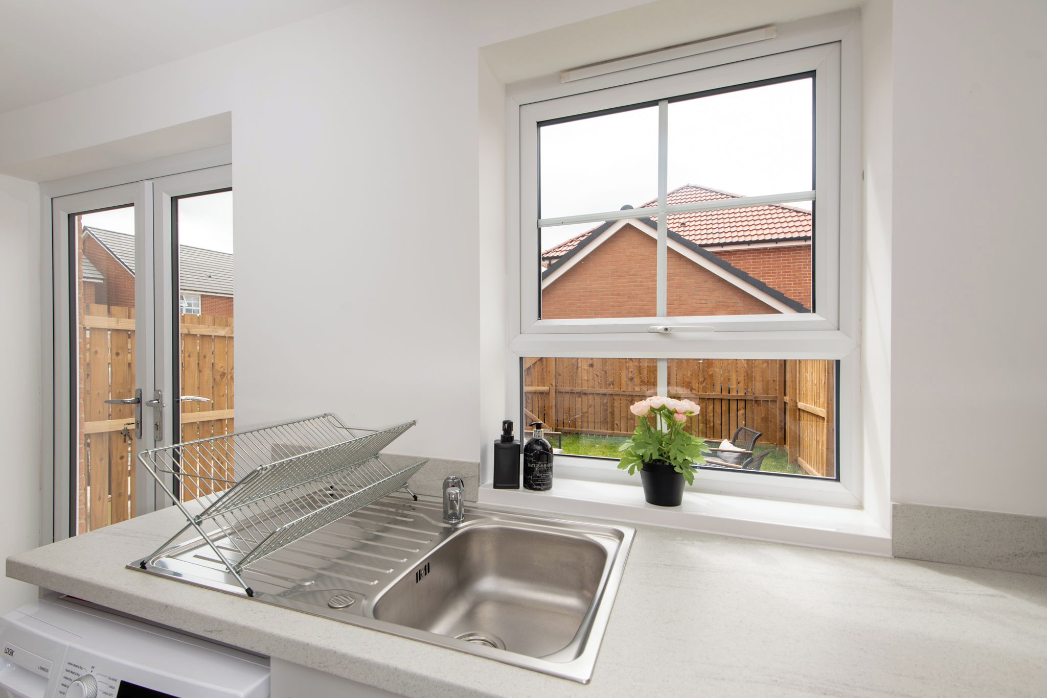 A kitchen with a stainless steel sink, a dish rack, a window with a view of a brick house and a wooden fence, and a potted plant on the windowsill.