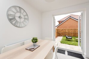 A room with a wooden table, a clock, and a view of a backyard with a fence and a bench.
