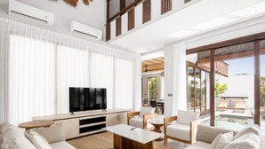 A modern living room with a flat-screen TV, a wooden coffee table, and a large sofa. There is a dining area with a table and chairs, and a glass-enclosed outdoor space with a pool. - 5-BR Beach Villa in Canggu Overlooking Rice Fields (Kuta Utara)