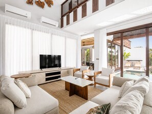 Un salon moderne avec un téléviseur à écran plat, une table basse en bois et un grand canapé. On y trouve également un coin repas avec une table et des chaises, ainsi qu’un espace extérieur clos par des baies vitrées, avec une piscine.