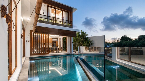 A modern two-story house with a glass-enclosed balcony, a swimming pool, and a well-lit interior. - 5-BR Beach Villa in Canggu Overlooking Rice Fields (Kuta Utara)