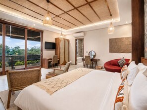A bedroom with a large bed, two chairs, a desk, a TV, and a view of greenery.