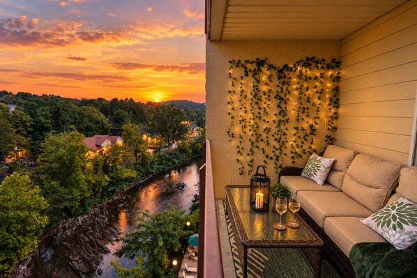 A balcony with a sofa, a table with glasses and a candle, and a view of a river and sunset.