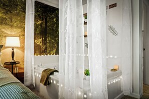 A bedroom with a bed, a bedside lamp, a window with a view of trees, a bathtub, and a shower area with a curtain.