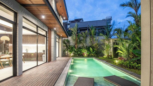 Uma área de piscina moderna ao ar livre, com deck de madeira, espreguiçadeiras e um alpendre envidraçado.
