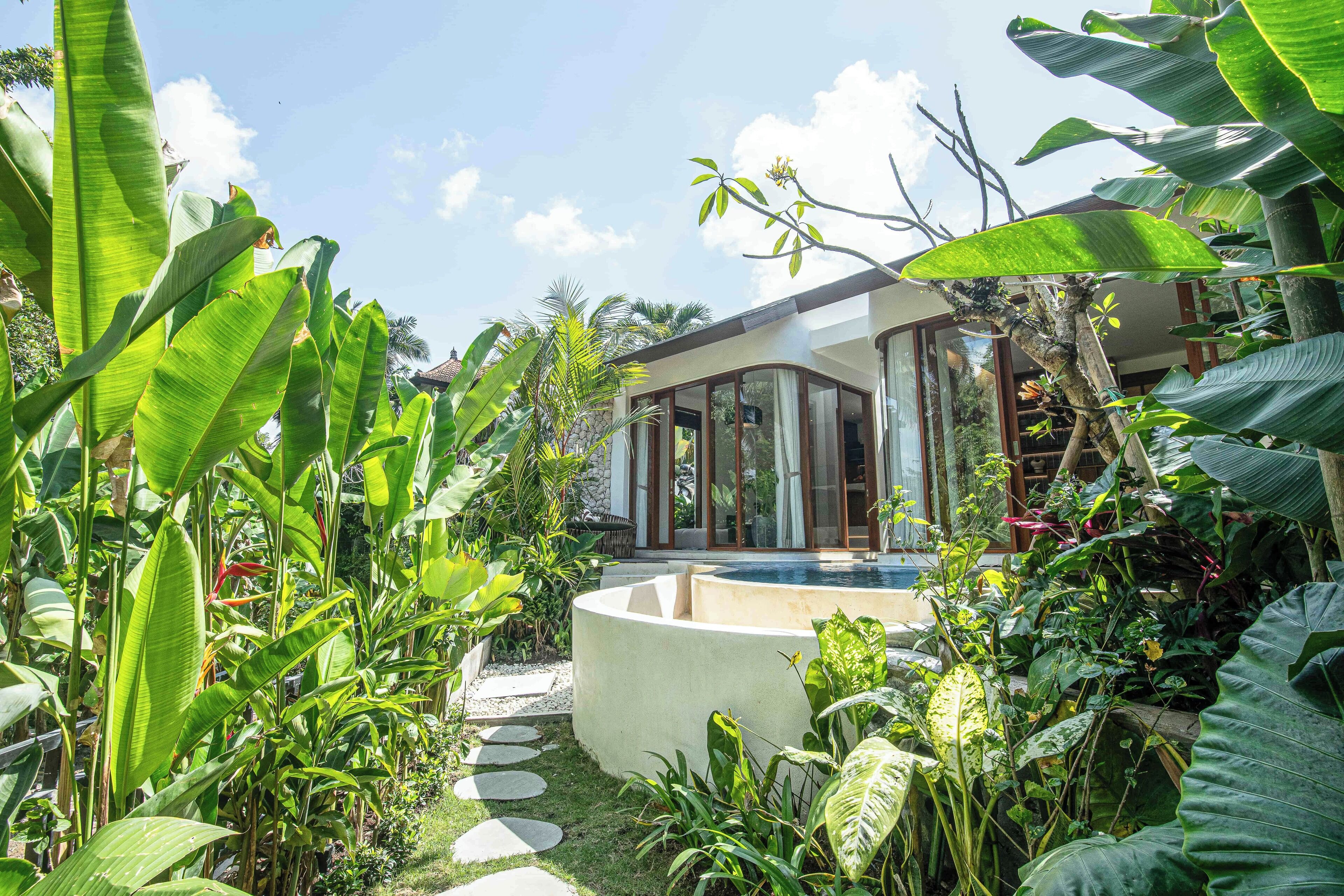 A modern house with a glass facade, surrounded by lush tropical plants and a small circular water feature.