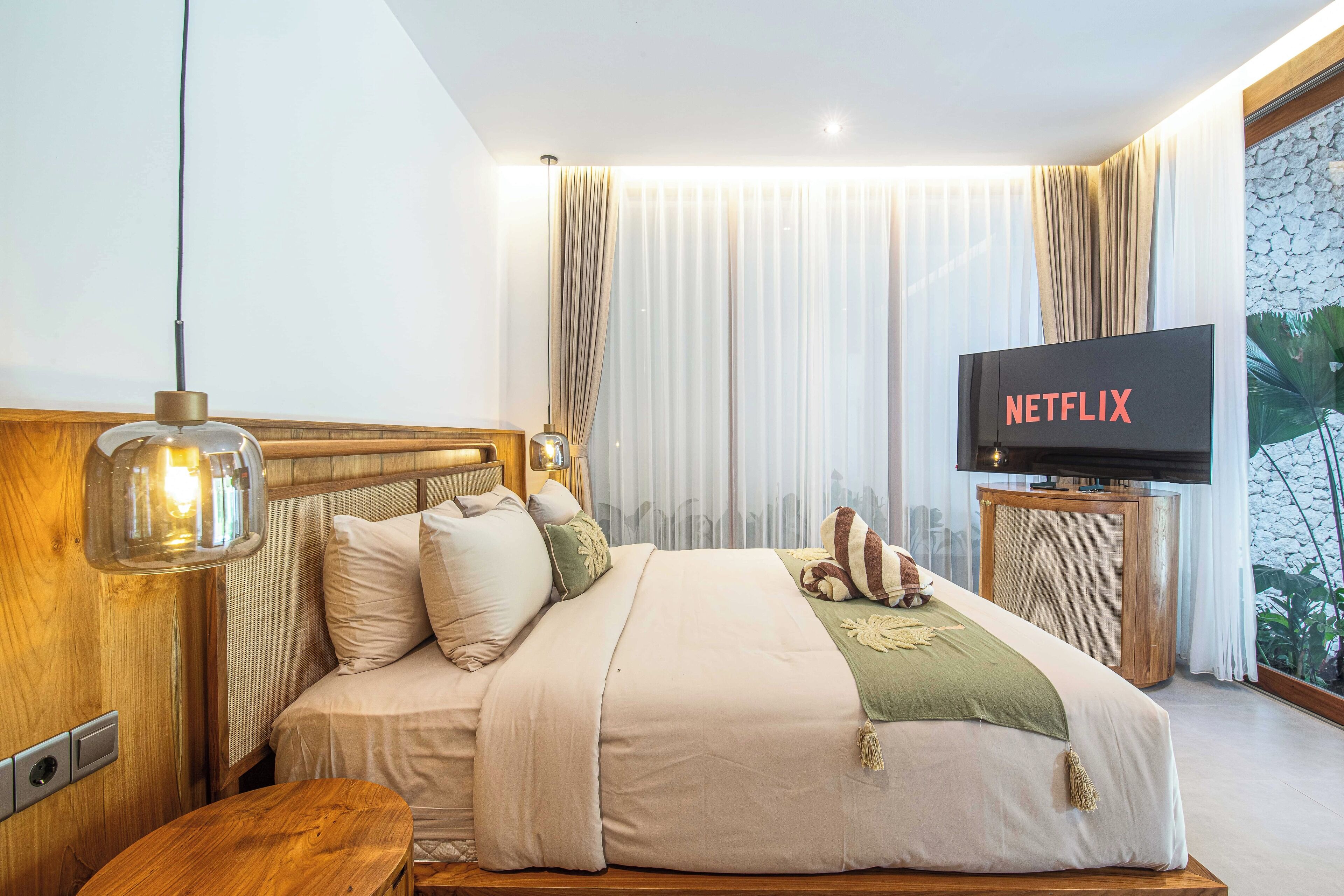 A modern hotel room with a large bed, a wooden headboard, a wooden nightstand, a television on a stand, and a large window with curtains.