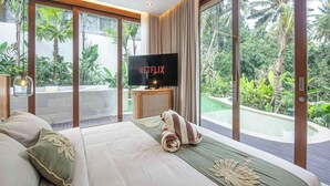 Un dormitorio con una cama grande, televisor y vistas a una piscina y plantas tropicales.