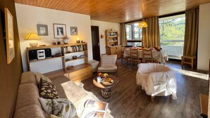 Uma sala de estar aconchegante com teto de madeira, um sofá, uma mesa de centro com frutas, uma estante de livros e uma área de refeições com mesa e cadeiras.
