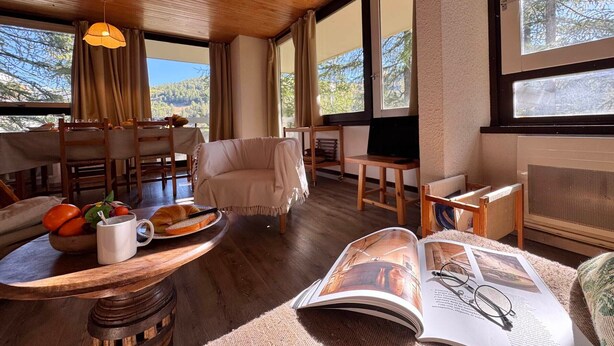 Uma sala de estar acolhedora com teto de madeira, janelas amplas, área de jantar com mesa posta para dois e um ambiente de estar confortável.