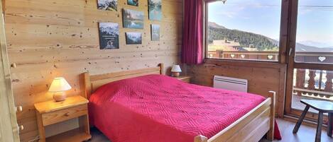 A wooden room with a bed, a nightstand, a lamp, and a window with a view of mountains.