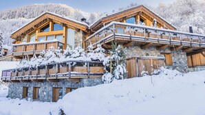 Ein Chalet mit mehreren Balkonen, Steinwänden und Holzwerteln, umgeben von schneebedeckten Bäumen.