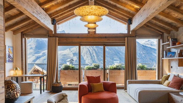 Ein geräumiges Wohnzimmer mit großem Fenster und Blick auf die Berge, Holzbalken an der Decke sowie einer gemütlichen Sitzecke mit einem roten Sessel und einem grauen Sofa.