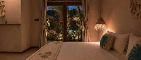 A bedroom with a bed, a nightstand, a macrame wall hanging, and a view of a garden through a glass door.