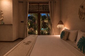 A bedroom with a bed, a nightstand, a macrame wall hanging, and a view of a garden through a glass door.