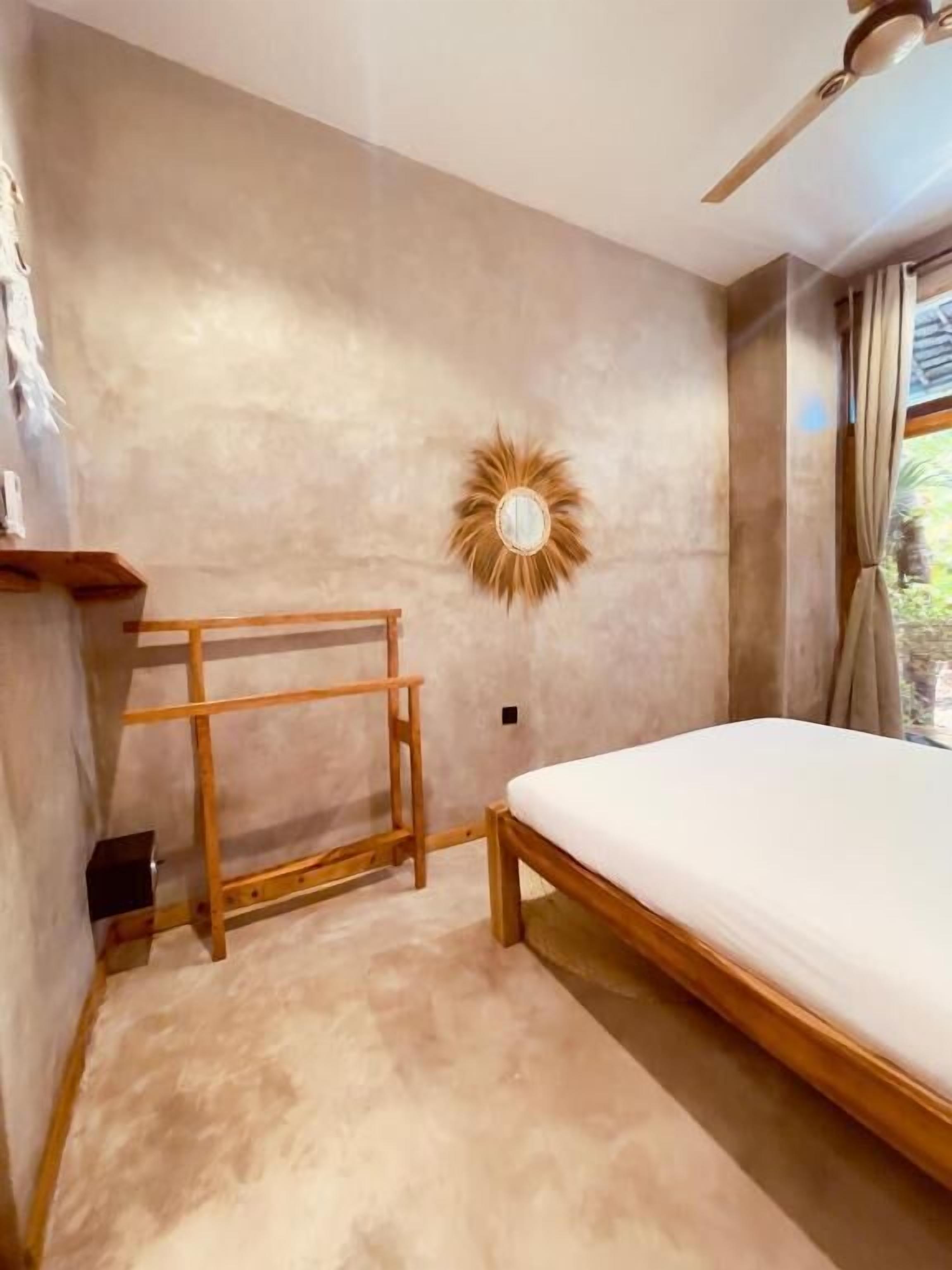 Un dormitorio con una cama, un banco de madera, un espejo montado en la pared y un ventilador de techo.