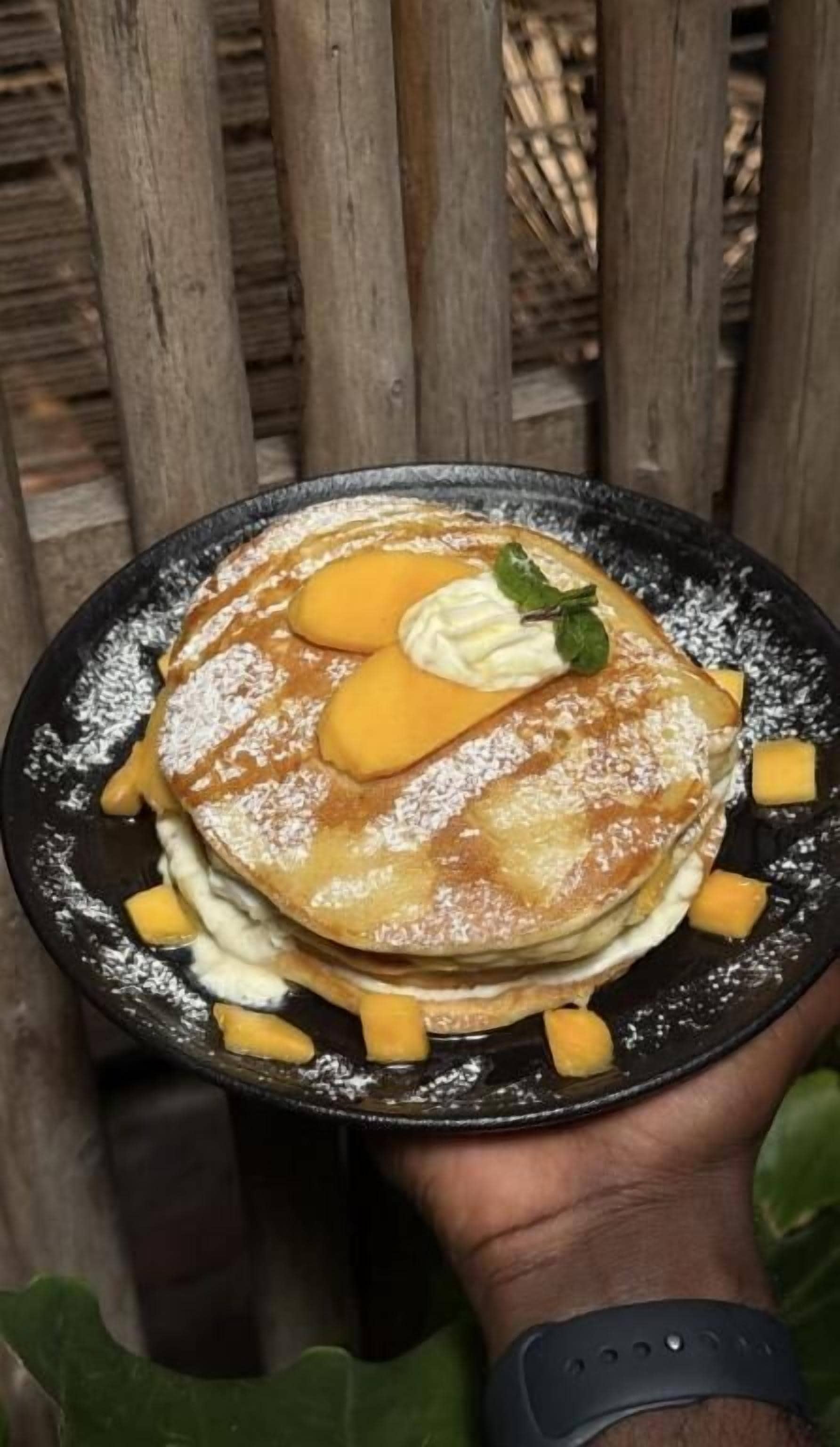 A stack of pancakes with syrup, whipped cream, and a mint leaf on a black plate.