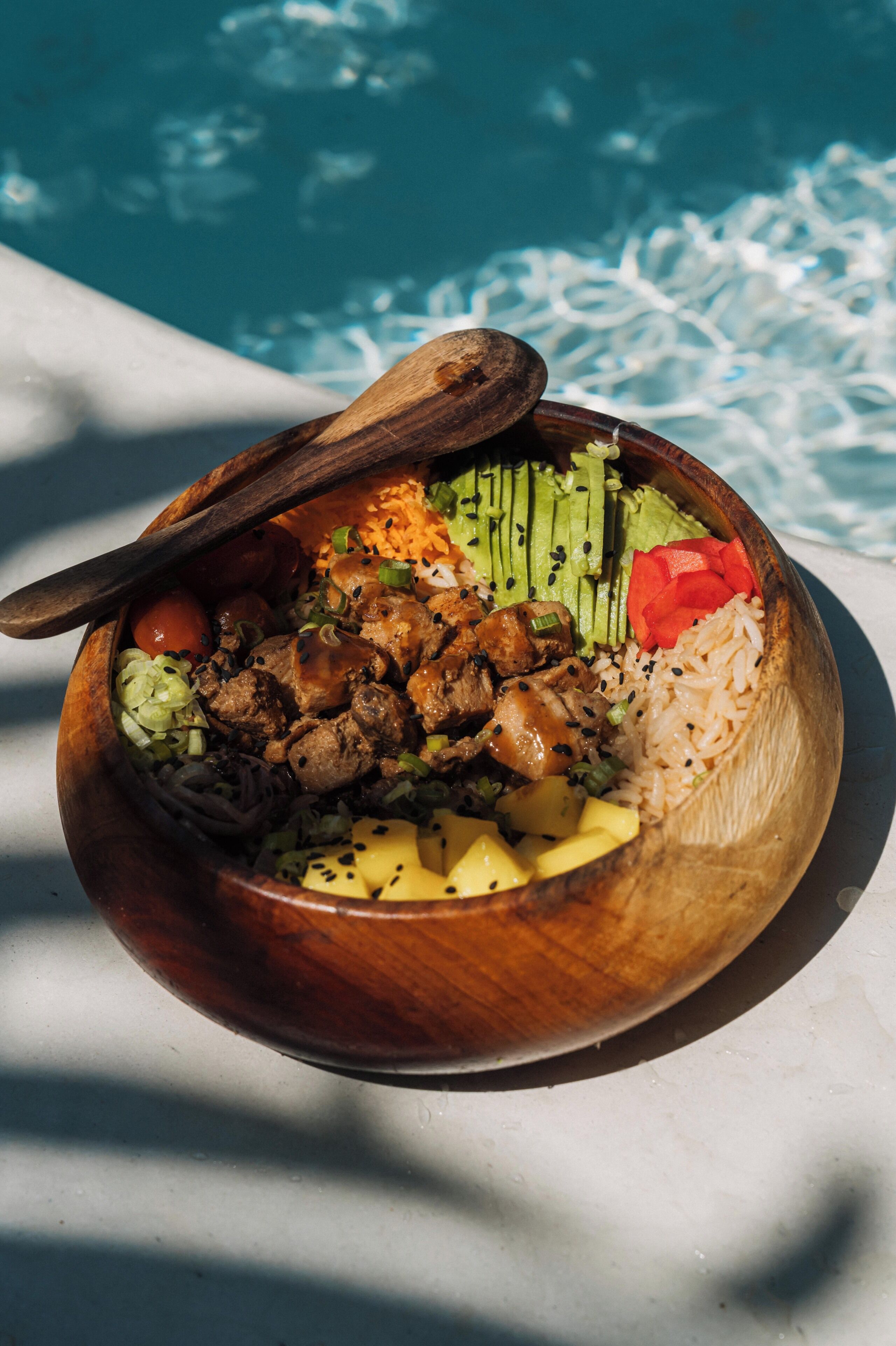 A wooden bowl filled with a variety of foods placed on a surface with a pool in the background.
