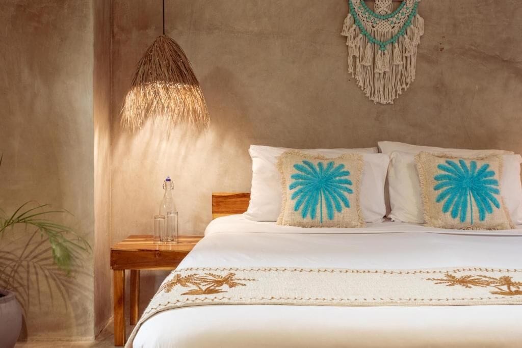 A bed with white bedding and blue floral pillows, a wooden nightstand with a bottle, a macramé wall hanging, and a hanging light fixture.