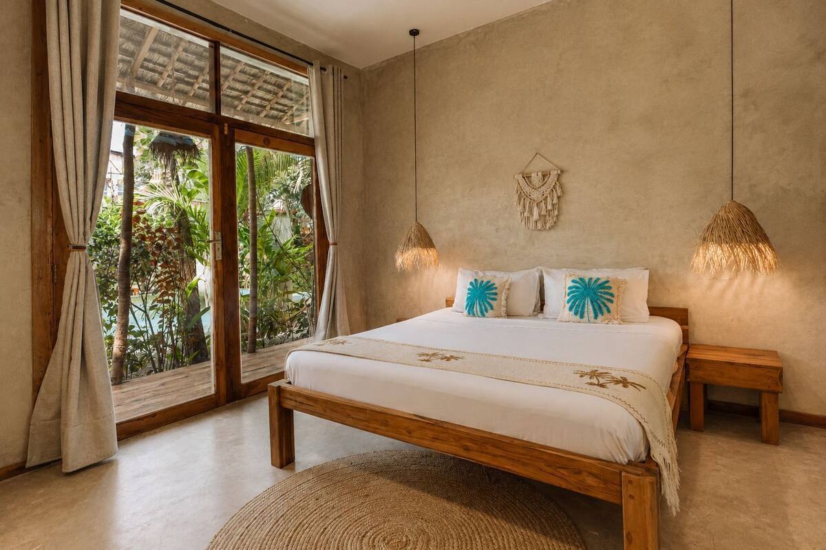 Ein Schlafzimmer mit einem hölzernen Bett, zwei Nachttischen, einem gewebten Teppich, einem großen Fenster mit Vorhängen und einer Wanddeko aus Makramee.