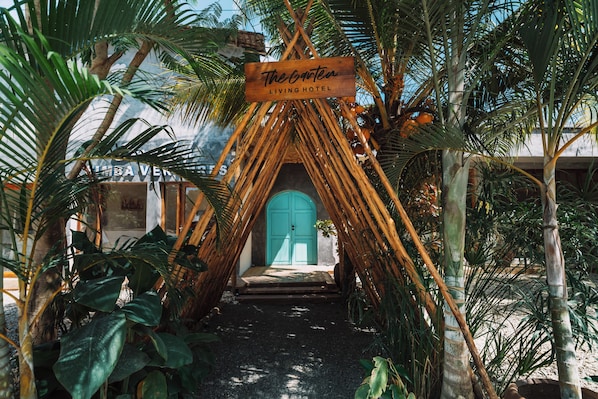 A tropical entrance with a wooden sign reading 'The Orenot Living Hotel,' surrounded by lush greenery and a pathway leading to a turquoise door.