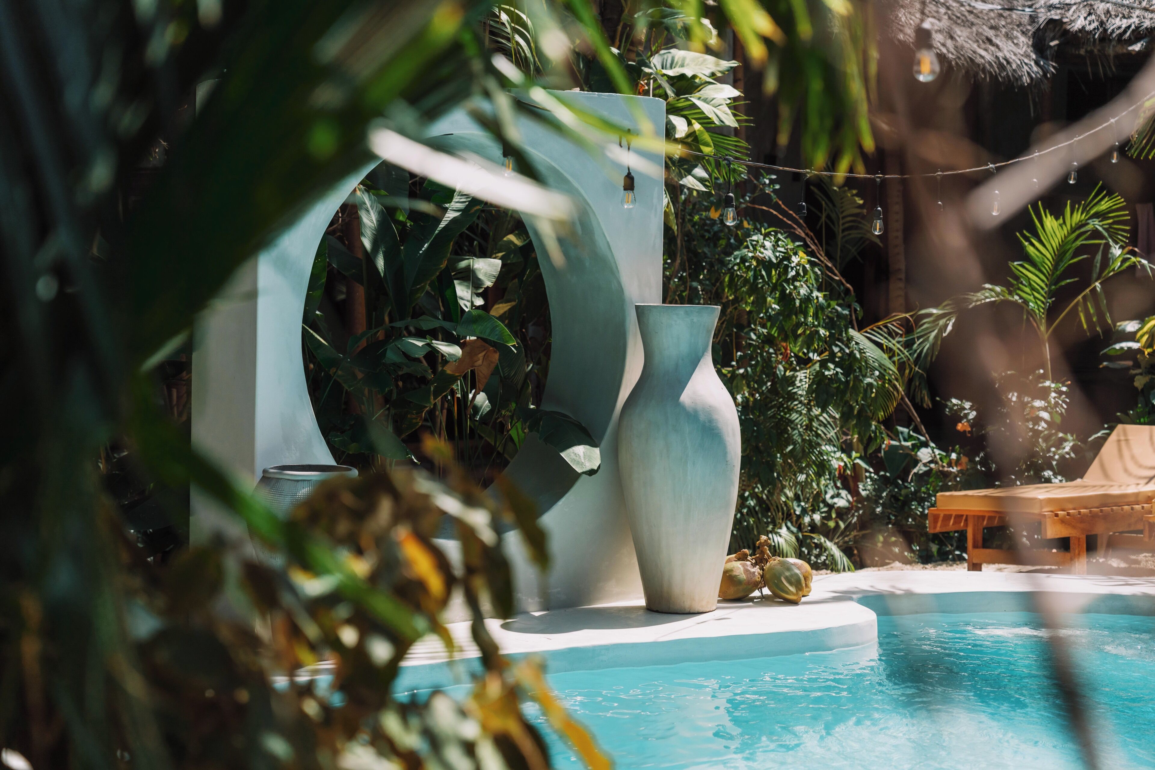A pool area with a large vase, a wooden bench, and lush greenery.